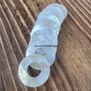 May include: A collection of iridescent, round, mother-of-pearl beads with a central hole. The beads are arranged on a wooden surface. The text "LoveCraftBeads" is visible on the image.