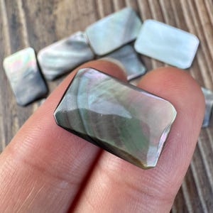 May include: A rectangular, faceted mother-of-pearl cabochon with a smooth, polished surface. The piece displays iridescent hues of gray, green, and soft pink, reflecting light across its beveled edges. Several similar pieces are visible in the background.