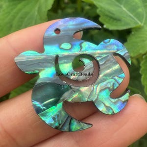 May include: Abalone shell pendant in a stylized turtle shape. The iridescent surface displays shades of blue, green, and purple. The pendant has a spiral design and the text "LoveCraftBeads".