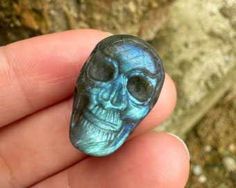 Puede incluir: Un cristal de labradorita azul tallado en forma de cráneo. El cristal tiene un brillo iridiscente.