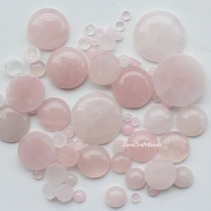 May include: A collection of pink rose quartz cabochons in various sizes. The cabochons are smooth and polished, with a light pink colour. The cabochons are arranged in a random pattern on a white background. LoveCraftBeads is written in the bottom right corner of the image.