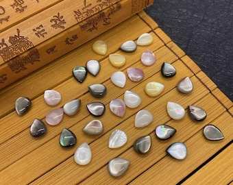 May include: A collection of iridescent, teardrop-shaped shell cabochons in various shades of white, grey, and pink. The cabochons are arranged on a bamboo surface.