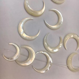 May include: A collection of iridescent, crescent-shaped beads. The beads are a creamy white color and have a pearlescent sheen. They are strung together on a clear string, suggesting they are intended for jewelry making or other craft projects.