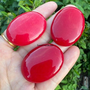 Può includere: Tre pietre rosse lisce, levigate e di forma ovale con una finitura lucida. Le pietre sono di un colore rosso vibrante e hanno un aspetto uniforme e arrotondato. Il testo LoveCraftBeads è visibile in lettere nere sulla pietra centrale.