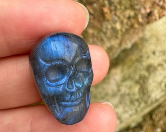 Puede incluir: Un cráneo tallado en un cristal de labradorita azul. El cristal tiene un color azul oscuro con destellos iridiscentes de verde y dorado.