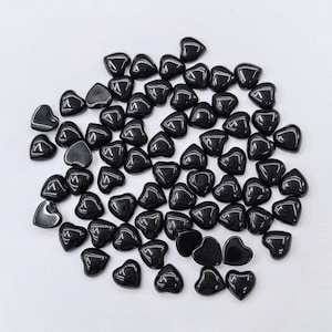 May include: A collection of numerous small, polished black heart-shaped beads. Each bead features a smooth, glossy surface with a reflective finish, arranged in a loose, scattered cluster across the frame, showcasing uniform size and shape.
