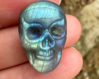 Puede incluir: Un cráneo tallado en un cristal de labradorita azul y verde. El cristal tiene un brillo iridiscente.