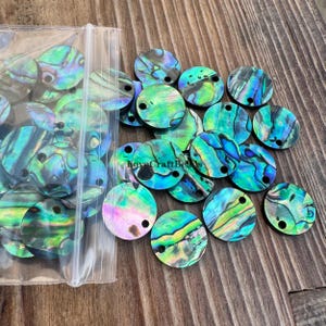 Peut inclure: Collection de perles rondes en coquillage d'ormeau irisé, percées d'un trou. Les perles présentent des couleurs variées, notamment des bleus, des verts et des violets. Certaines perles sont en vrac, d'autres dans un sac en plastique transparent. Le texte "LoveCraftBeads" est visible.