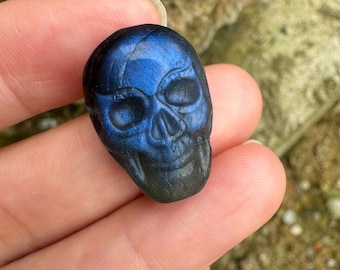 Puede incluir: Una talla de calavera de labradorita azul y negra. La calavera está detallada con cuencas de los ojos, una nariz y una boca.