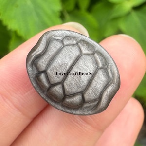 Puede incluir: Una cuenta de caparazón de tortuga gris metálico con una superficie texturizada. La cuenta tiene un patrón segmentado y el texto "LoveCraftBeads" es visible. La cuenta se sostiene en una mano sobre un fondo verde borroso.