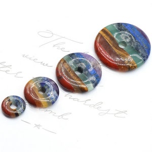 Op de afbeelding: Vier chakra-steen donut-hangers in verschillende maten. De hangers zijn gemaakt van zeven verschillende stenen, die elk een andere chakra vertegenwoordigen. De stenen zijn gerangschikt in een cirkel met een gat in het midden. De stenen zijn rood, oranje, geel, groen, blauw, indigo en violet.