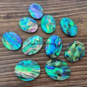 Puede incluir: Diez cabujones de concha de abulón de forma ovalada con remolinos iridiscentes en tonos vibrantes de verde, azul y morado. Cada pieza muestra un patrón nacarado natural único con una superficie lisa y pulida. El texto LoveCraftBeads es visible.