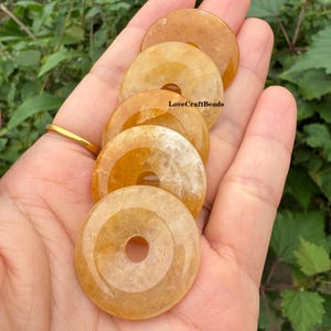 May include: A hand holds several golden-yellow gemstone donut beads. Each bead has a central hole and concentric circle design. The beads are smooth and translucent, with the text "LoveCraftBeads" visible on one of the beads.