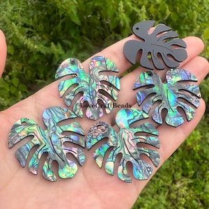 Puede incluir: Una colección de colgantes con forma de hoja en varios colores. Los colgantes tienen un acabado veteado e iridiscente con tonos verdes, azules y morados. Un colgante es negro liso. Los colgantes se sostienen en una mano sobre un fondo verde.