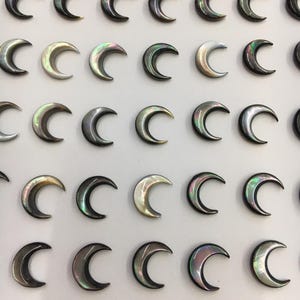 Op de afbeelding: Een reeks iriserende, halvemaanvormige hangers. De hangers hebben een parelmoerglans met kleuren variërend van zilver tot zwart, met hints van groen en goud. De manen zijn gerangschikt in een rasterpatroon tegen een witte achtergrond.