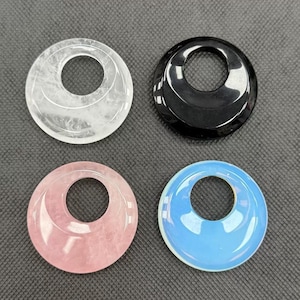 Puede incluir: Cuatro colgantes circulares con un agujero en el centro. Los colgantes son transparentes, negros, rosas y azules. El colgante transparente tiene una superficie texturizada. El colgante rosa es cuarzo rosa. El colgante azul tiene un aspecto opalescente.