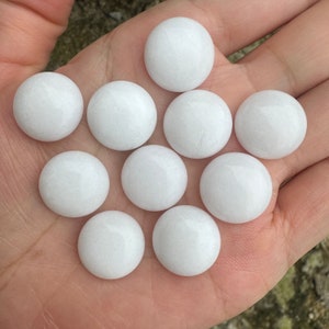 Puede incluir: Diez piedras blancas, redondas, lisas y pulidas. Las piedras son aproximadamente del mismo tamaño y están dispuestas en semicírculo sobre una mano.