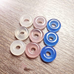 Könnte beinhalten: Neun durchscheinende, donut-förmige Perlen in Weiß, Rosa und Blau. Die Perlen sind in einem Raster angeordnet, jede mit einem zentralen Loch. Die Perlen sind glatt und glänzend und reflektieren das Licht.