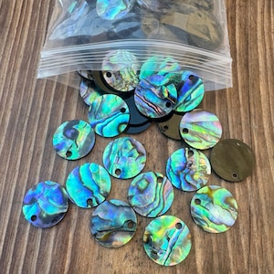 Könnte beinhalten: Eine Sammlung schillernder, runder Abalone-Muschelkernperlen mit einem Loch durch jede. Die Perlen zeigen eine Reihe von Farben, darunter Blau-, Grün- und Lilatöne, und sind auf einer Holzoberfläche verstreut.