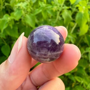 May include: A smooth, polished amethyst sphere with a deep purple hue and white striations. The sphere is held in a hand, showcasing its size and shape.