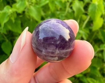 May include: A smooth, polished amethyst sphere with a deep purple hue and white striations. The sphere is held in a hand, showcasing its size and shape.