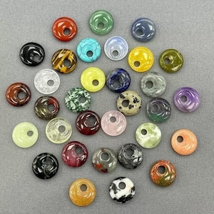 Könnte beinhalten: Eine Sammlung farbenfroher Donut-Perlen aus Edelsteinen in verschiedenen Farben, darunter Rot, Grün, Blau, Gelb und Lila. Jede Perle hat ein Loch in der Mitte, perfekt für die Schmuckherstellung. Die Perlen sind auf einer grauen Oberfläche angeordnet.