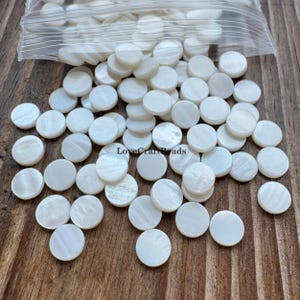 May include: A collection of round, white mother-of-pearl beads scattered on a wooden surface. The beads are in various sizes and shapes, with a pearlescent sheen. The text "LoveCraftBeads" is visible on the surface.