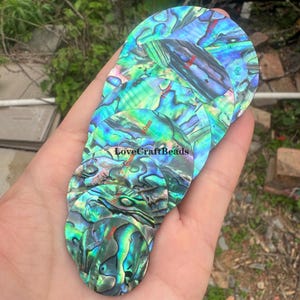 Puede incluir: Una colección de piezas ovaladas e iridiscentes de concha de abulón en tonos verdes, azules y morados. Las piezas varían en tamaño, con medidas en mm visibles. También se ve el texto "LoveCraftBeads".