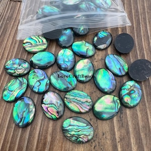 May include: Oval-shaped abalone shell cabochons in a variety of iridescent colors, including green, blue, and purple. Some cabochons are loose, while others are in a clear plastic bag. The text "LoveCraftBeads" is visible.