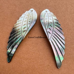May include: Two iridescent, carved mother-of-pearl wing pendants. Each wing features a detailed feather design with a hole at the top for stringing. The wings have a pearlescent sheen with hints of green, purple, and black. The text "LoveCraftBeads" is visible.