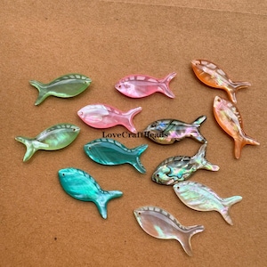 Könnte beinhalten: Sortiment von 10 regenbogenfarbenen Fisch-Anhängern aus Muschelschalen in verschiedenen Farben, darunter Rosa, Grün, Blau und Weiß. Die Anhänger sind aus Perlmutt und haben ein natürliches, organisches Aussehen.