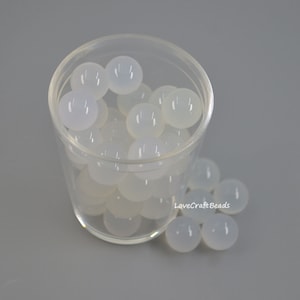 May include: A clear plastic container filled with white round beads. The beads are scattered on a gray surface. LoveCraftBeads is written on the surface.