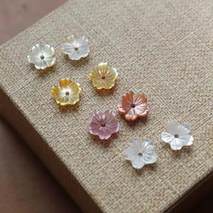 Può includere: Una collezione di perline a forma di fiore in vari colori, tra cui bianco, giallo, rosa e arancione. Le perline hanno una lucentezza perlescente e sono disposte su una superficie strutturata color beige. Ideale per la creazione di gioielli.