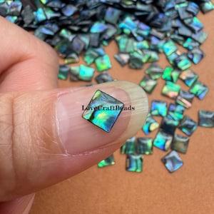 Könnte beinhalten: Nahaufnahme einer quadratischen, schillernden Perle, die von einer Person gehalten wird. Die Perle hat eine Mischung aus blauen, grünen und violetten Farbtönen. Der Text "LoveCraftBeads" ist sichtbar. Viele ähnliche Perlen sind im Hintergrund verstreut.