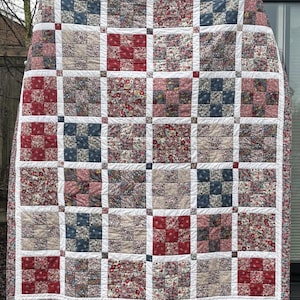 May include: A patchwork quilt with a floral and geometric design. The quilt is composed of squares in shades of red, blue, and neutral tones, separated by white borders. The quilt is displayed outdoors, highlighting its detailed construction and colour palette.