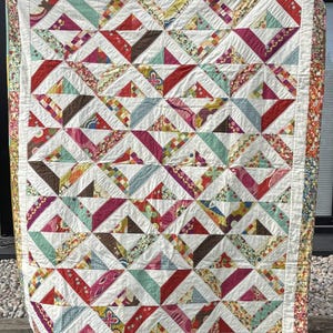 May include: A handmade quilt with a geometric pattern of colorful triangles and stripes. The quilt features a variety of fabrics in shades of red, pink, teal, brown, and floral prints, set against a white background. The border is a floral print.
