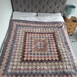 Könnte beinhalten: Eine handgefertigte Steppdecke mit einem Rautenmuster in Rosa-, Rot-, Grau- und Cremetönen bedeckt ein Bett. Das Bett hat ein graues, getuftetes Kopfteil und weiße Kissen. Ein Nachttisch aus Holz mit einer Lampe befindet sich neben dem Bett.