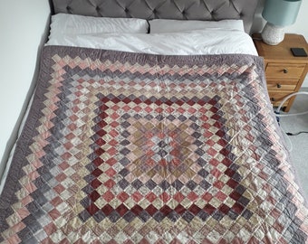 May include: A handmade quilt with a diamond pattern in shades of pink, red, gray, and cream covers a bed. The quilt is on a bed with a gray tufted headboard and white pillows. A wooden nightstand with a lamp is next to the bed.
