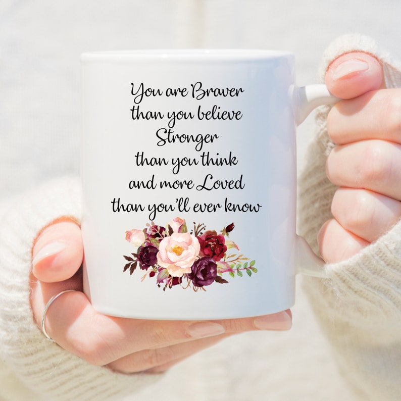 May include: White ceramic mug with a handle, featuring the text "You are Braver than you believe, Stronger than you think, and more Loved than you'll ever know." Adorned with a floral design in burgundy, pink, and green.