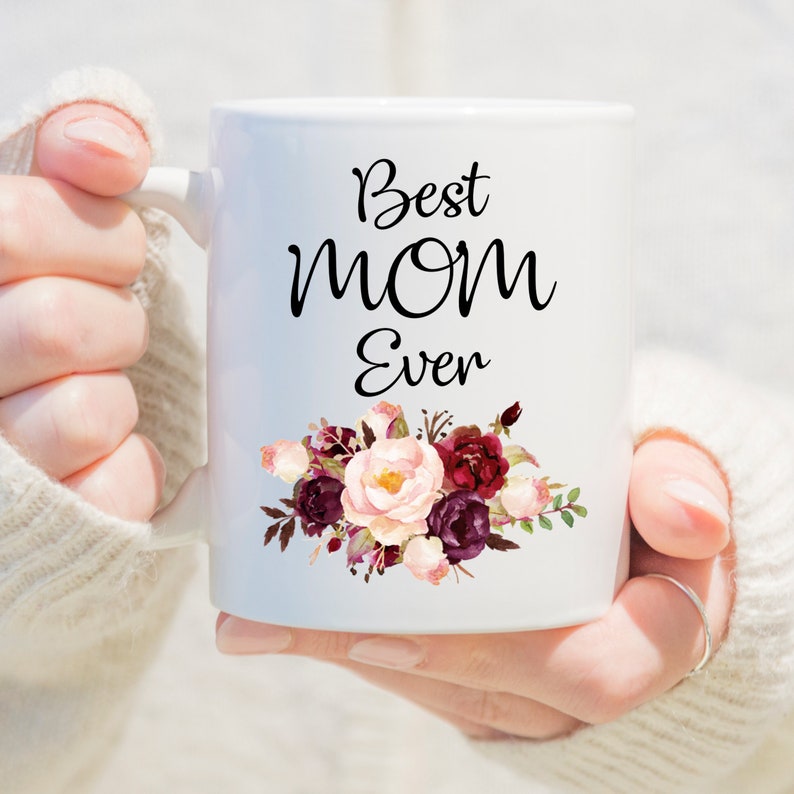 May include: White ceramic mug with the text "Best Mom Ever" in black script. The mug features a floral design in shades of pink, burgundy, and cream. The mug has a curved handle and is held by a person wearing a white sweater.