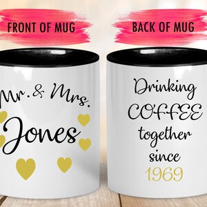 May include: Two white coffee mugs with black rims. The front mug has the text "Mr. & Mrs. Jones" with gold hearts. The back mug has the text "Drinking COFFEE together since 1969" with gold text.
