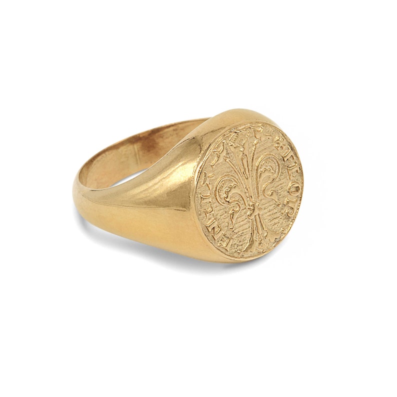 May include: A gold signet ring with a round face featuring an engraved fleur-de-lis design.
