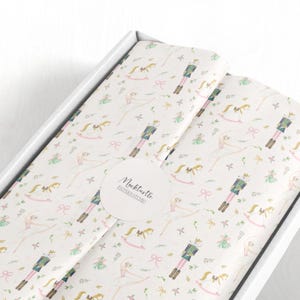 以下が含まれることがあります： バレリーナ、くるみ割り人形、ロッキングホース、リボンの繰り返し模様が施された白い薄葉紙。薄葉紙は白い箱の中に入っています。「Neckturtle Pattern Studio」と書かれた丸いステッカーが見えます。