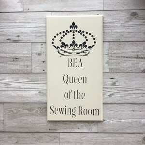 May include: A white wooden sign with black text and a black crown design. The sign reads "BEA Queen of the Sewing Room".