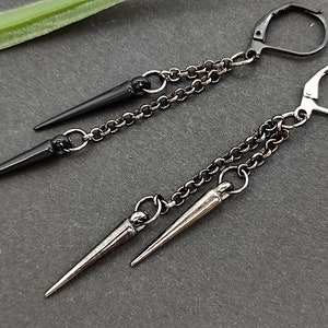 Op de afbeelding: Een paar oorbellen met zwarte en zilveren spikes die aan kettingen hangen. De oorbellen zijn gemaakt van metaal en hebben een eenvoudig ontwerp.