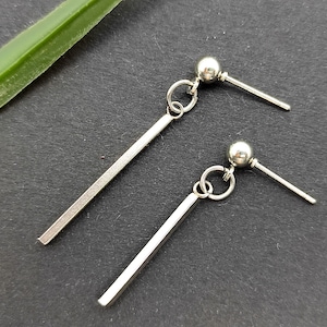 Puede incluir: Un par de pendientes plateados. Cada pendiente presenta un pequeño perno de bola, un anillo de conexión y una barra larga y rectangular. Los pendientes se muestran sobre una superficie gris oscuro, con una hoja verde en el fondo.