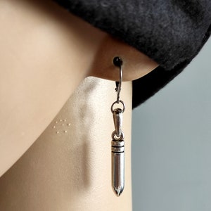 May include: Silver bullet shaped earrings hanging from a silver hook. The earrings are on a person's ear.