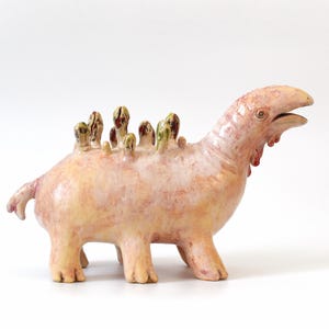 Può includere: Scultura in ceramica di una creatura fantastica, che combina elementi di dinosauro e uccello. Il corpo rosa presenta dettagli gialli e verdi, con una superficie strutturata. Poggia su quattro zampe, creando un pezzo decorativo unico.