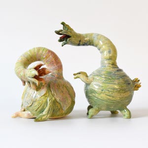 May include: Two ceramic sculptures in shades of green, yellow, and peach. One sculpture is curled with a floral-like form, the other has a bulbous body and a long, curved neck. Both have textured surfaces.