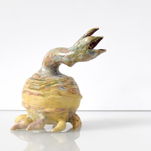 May include: A whimsical ceramic sculpture of a creature with a bulbous yellow body and a slender, curved neck. The sculpture has a textured surface with green, pink, and brown accents. The creature's head is open-mouthed, and it has small, stubby legs.